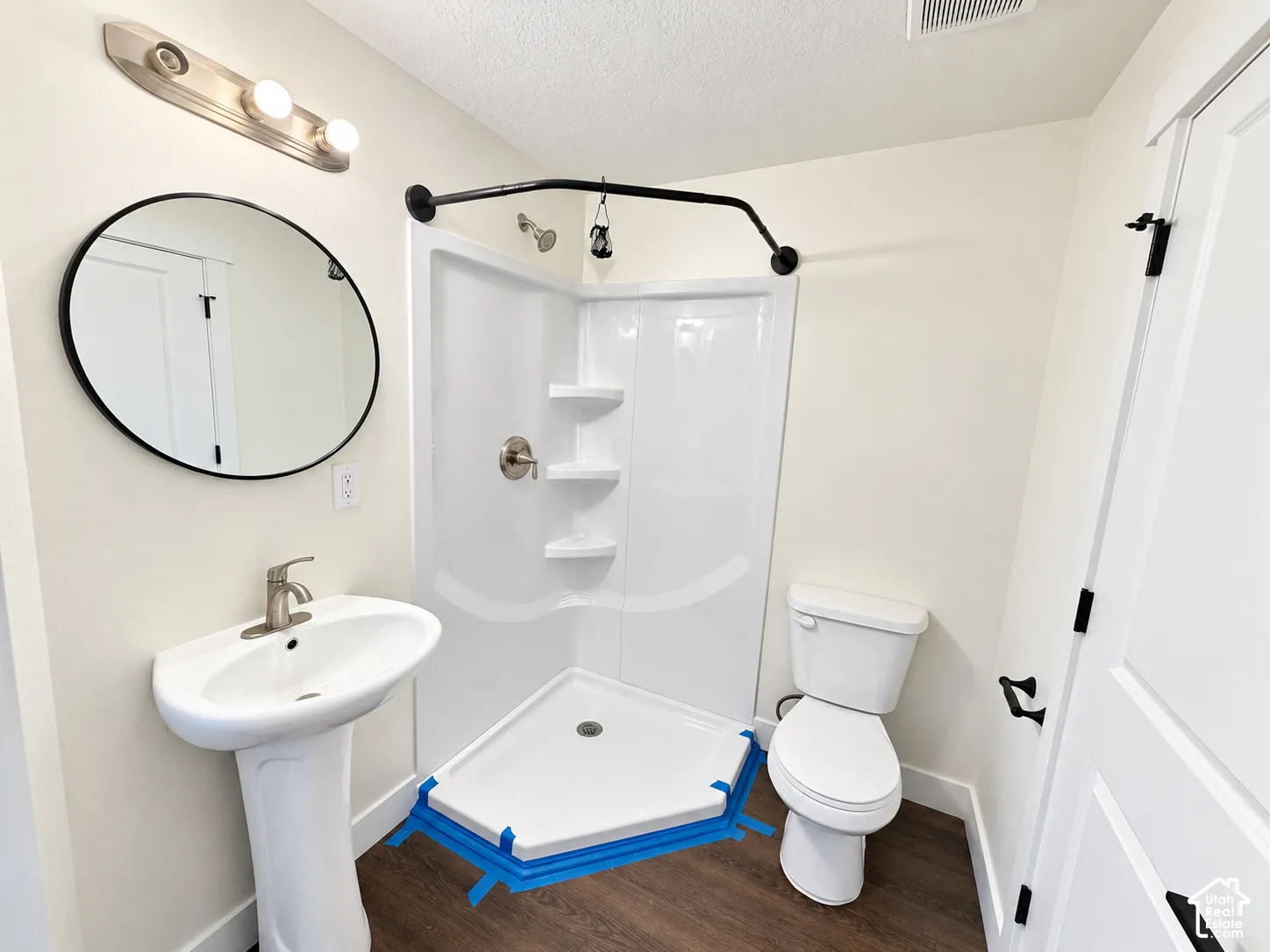 Pedestal sink with brushed nickel faucet, circular wall-mounted mirror, corner shower enclosure with integrated shelving, and wood-finish flooring