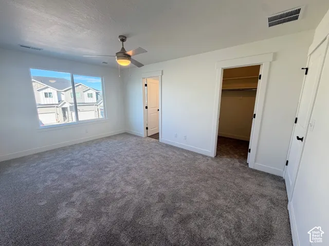 Neutral-toned wall-to-wall carpeting, ceiling fan with integrated lighting, oversized window, and walk-in closet with built-in shelving