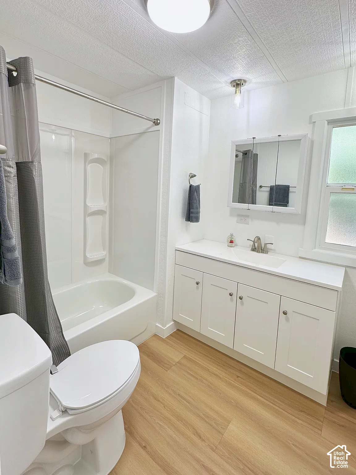 Bathroom featuring a white vanity cabinet with a solid surface countertop, an integrated sink, a multi-panel medicine cabinet, a tub-shower combination with built-in shelving, and wood-finish flooring