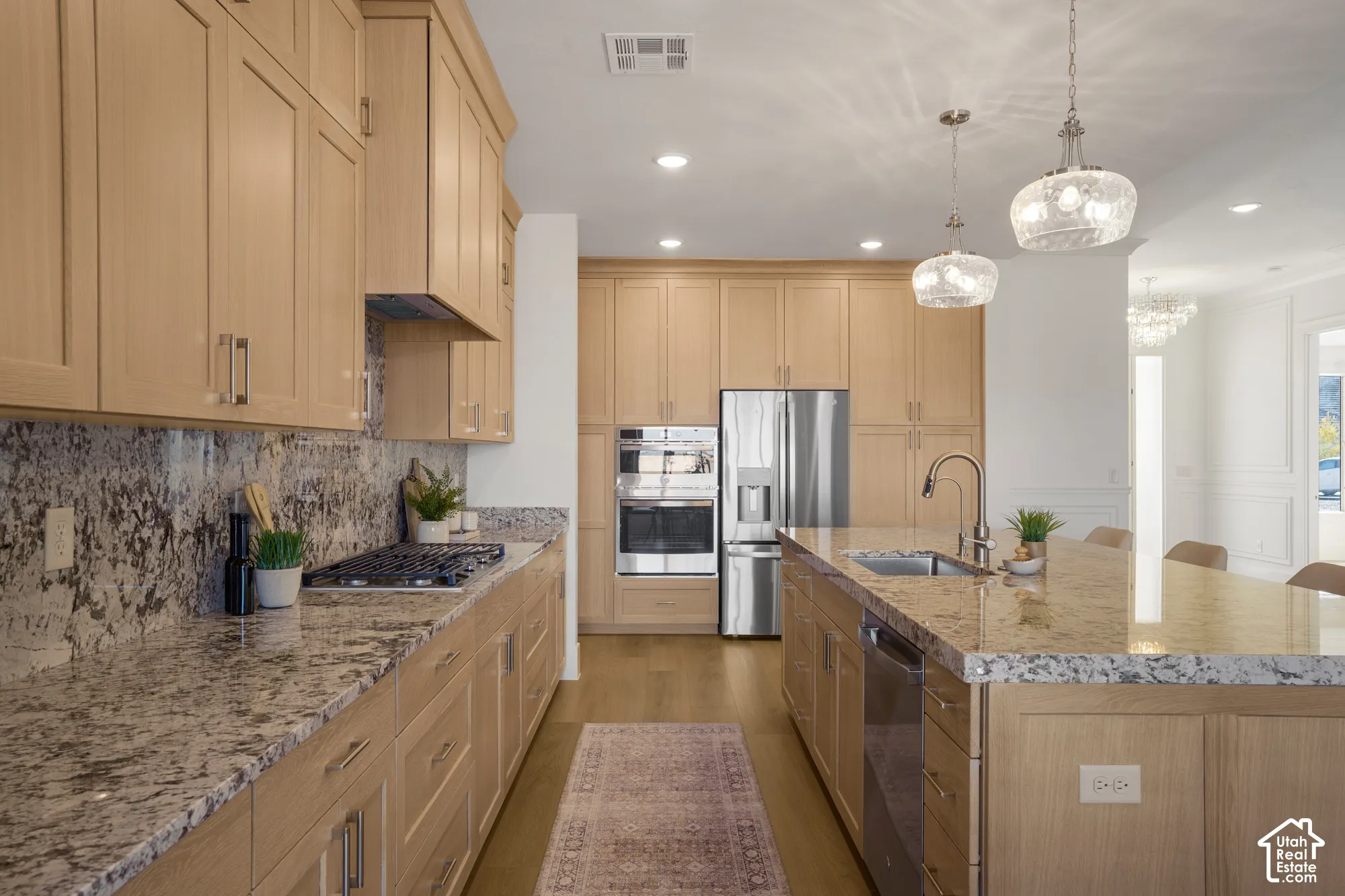 Kitchen featuring light wood cabinetry, stone countertops, stainless steel appliances, a central island with an undermount sink, and glass pendant lighting