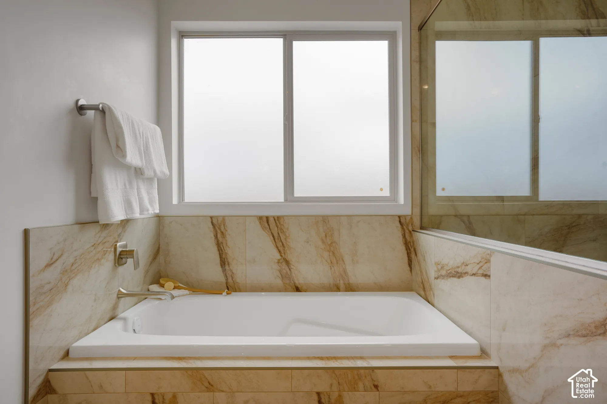 Soaking tub surrounded by stone-patterned tile, featuring brushed nickel plumbing fixtures, a sliding window, and a glass-enclosed shower