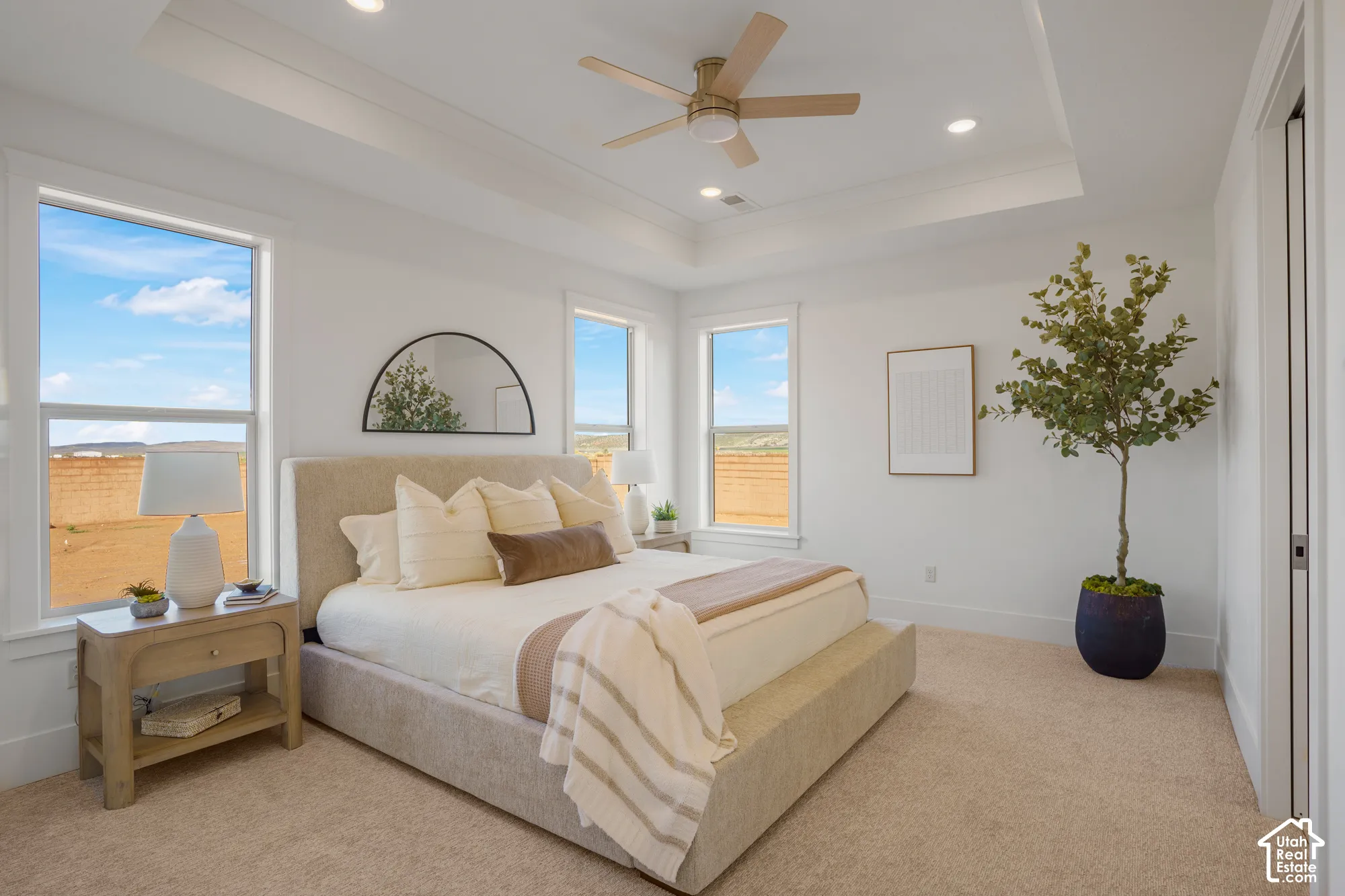 Tray ceiling with integrated recessed lighting, neutral carpeted flooring, three large windows, and a ceiling fan