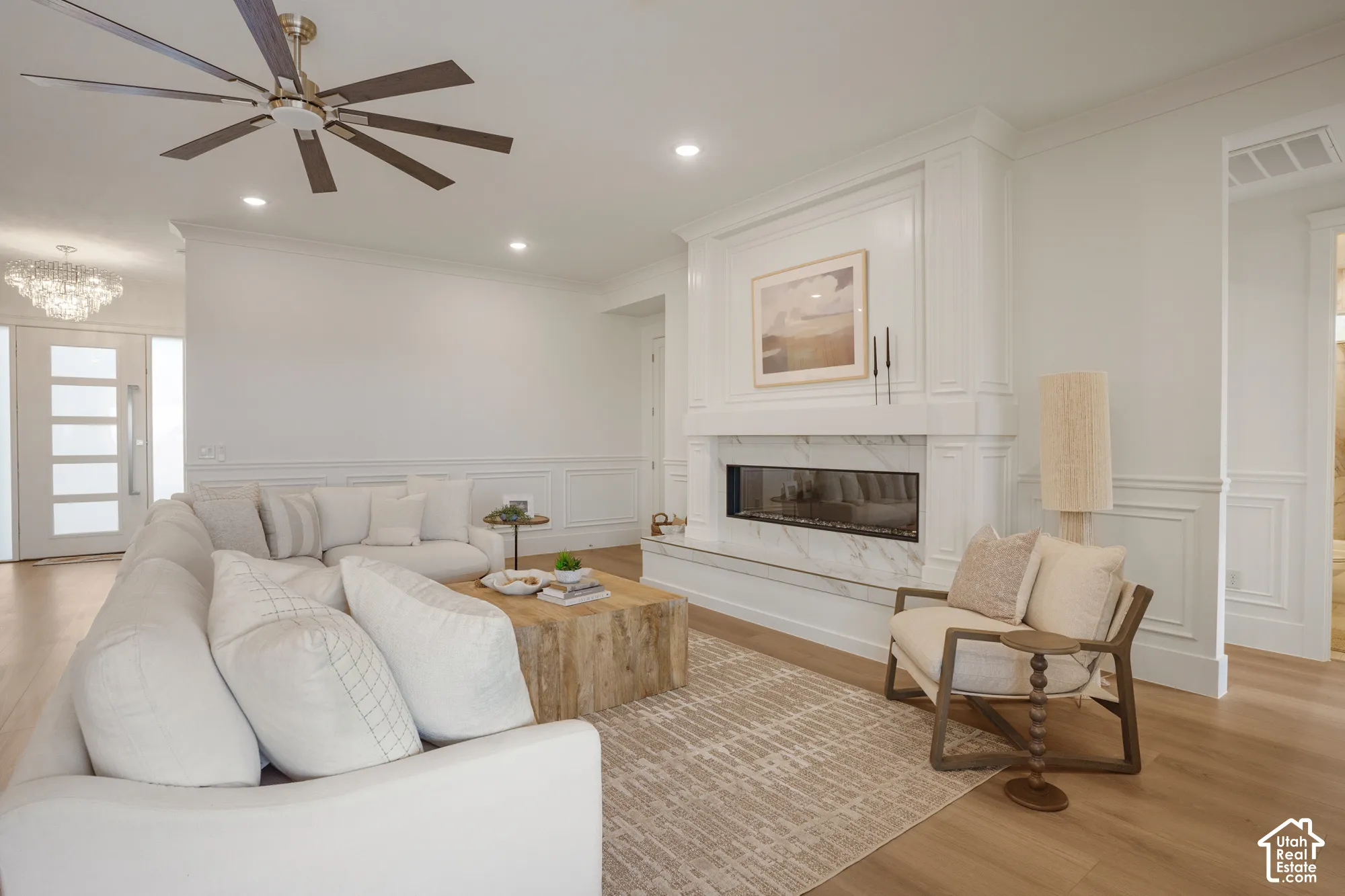 Linear fireplace with stone surround, custom millwork mantle, wood-finish flooring, wainscoting, and multi-blade ceiling fan