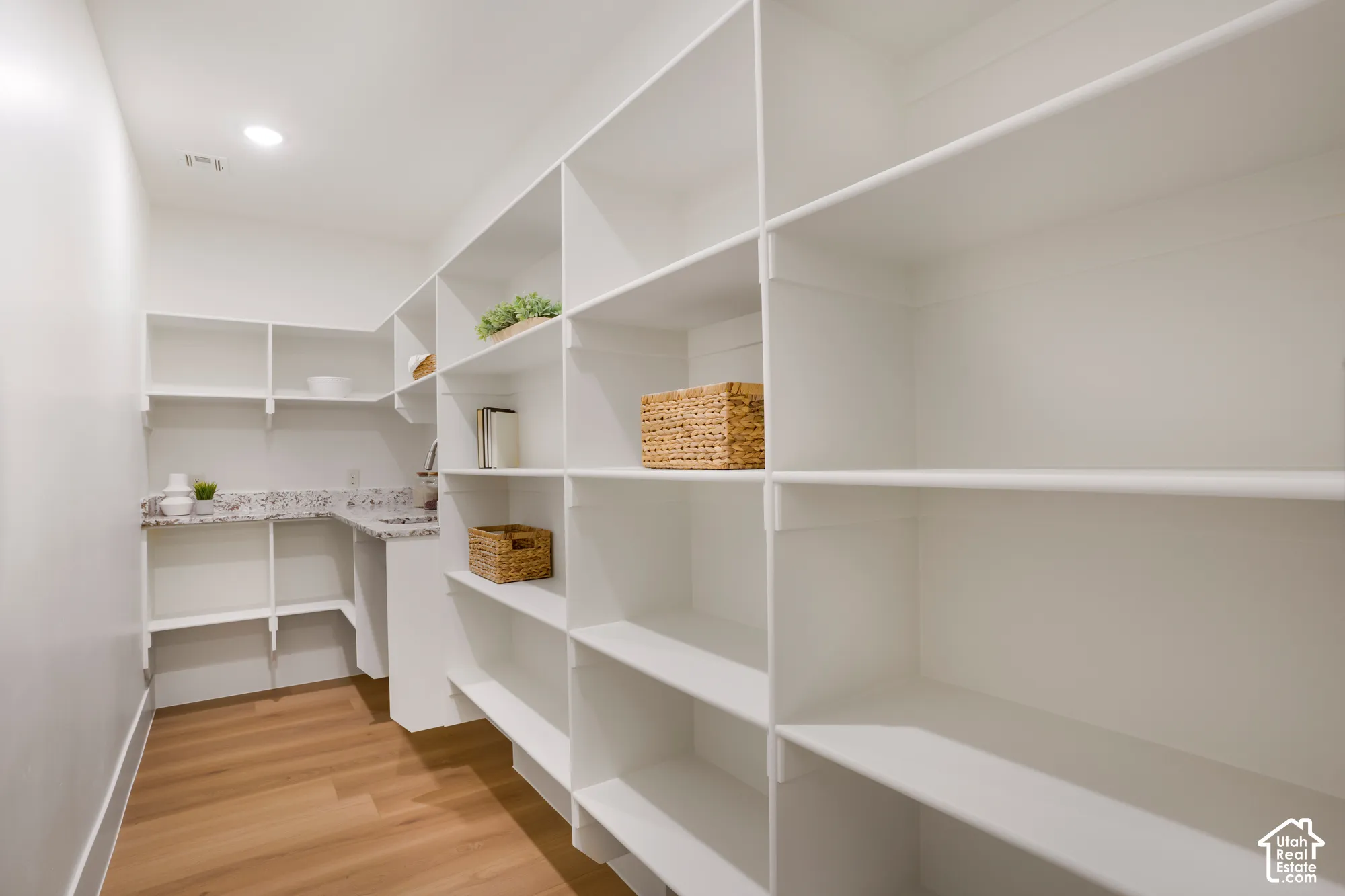 Walk-in storage space featuring extensive white built-in shelving, stone-finish countertops, wood-finish flooring, and recessed ceiling lighting
