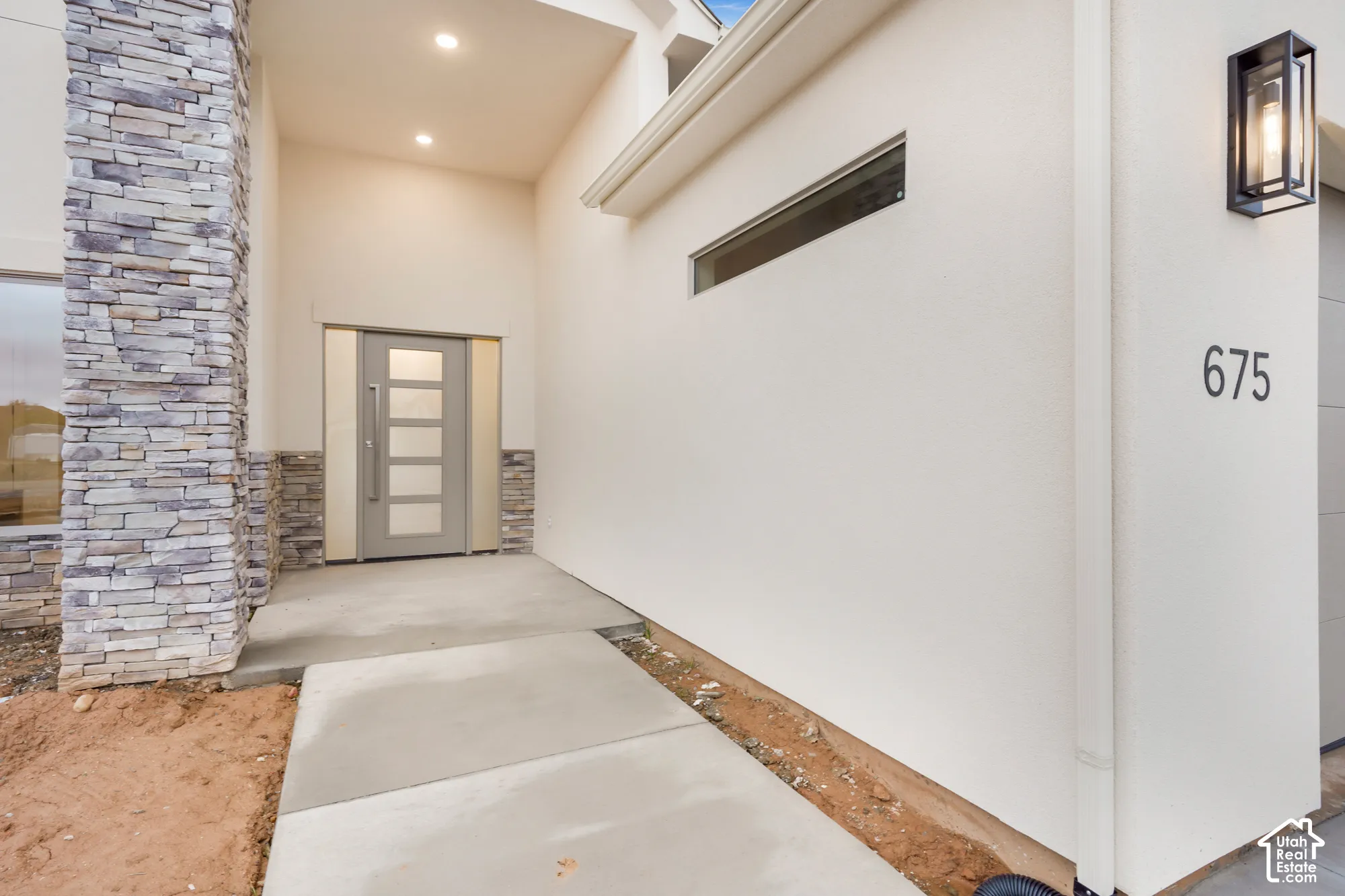 Modern entryway featuring a stacked stone facade, concrete walkway, solid-panel door with glass inserts, recessed lighting, and a geometric wall-mounted lantern