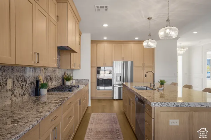Kitchen featuring light wood cabinetry, stone countertops, stainless steel appliances, a central island with an undermount sink, and glass pendant lighting