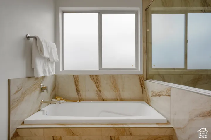 Soaking tub surrounded by stone-patterned tile, featuring brushed nickel plumbing fixtures, a sliding window, and a glass-enclosed shower