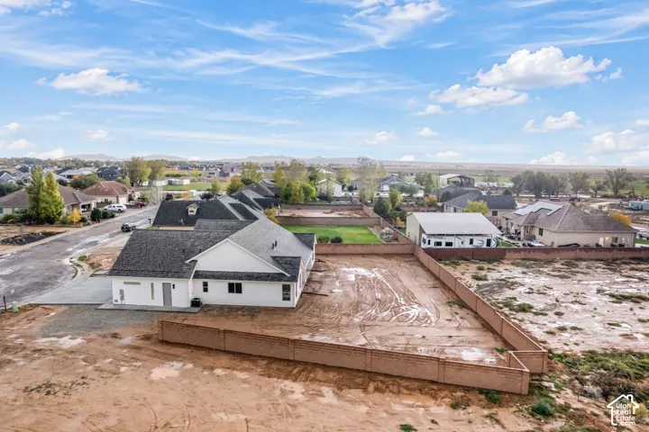 Aerial perspective of a single-story residence featuring a composite shingle roof, white exterior siding, a concrete driveway, and an expansive lot enclosed by a perimeter masonry wall