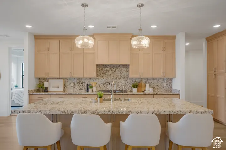 Kitchen featuring light-tone cabinetry, stone-finish countertops, a central island with integrated sink, stone-finish backsplash, and two hanging glass pendant lights