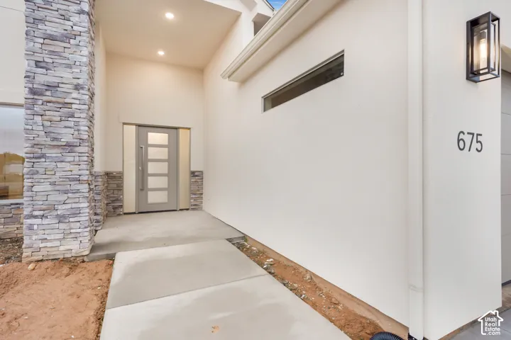 Modern entryway featuring a stacked stone facade, concrete walkway, solid-panel door with glass inserts, recessed lighting, and a geometric wall-mounted lantern