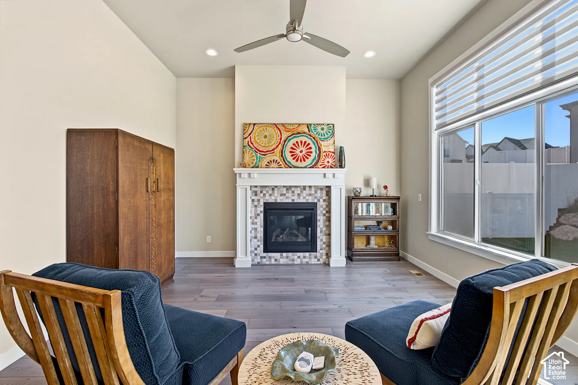 Fireplace with tile surround and white mantel, wood-finish flooring, ceiling fan, and large windows with horizontal blinds