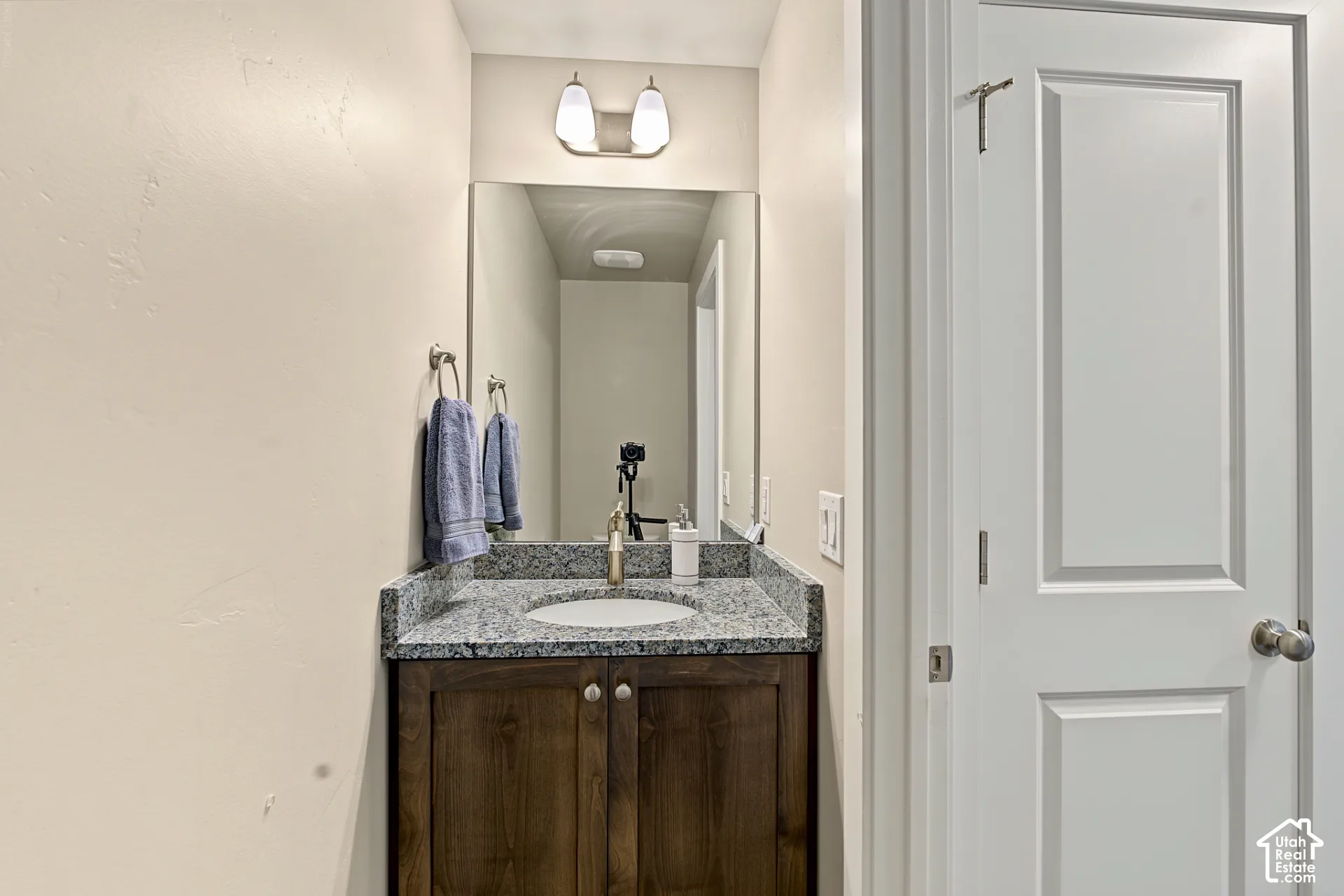 Vanity with stone countertop, undermount sink, dual-door wood-finish cabinet, wall-mounted mirror, and overhead dual-light fixture