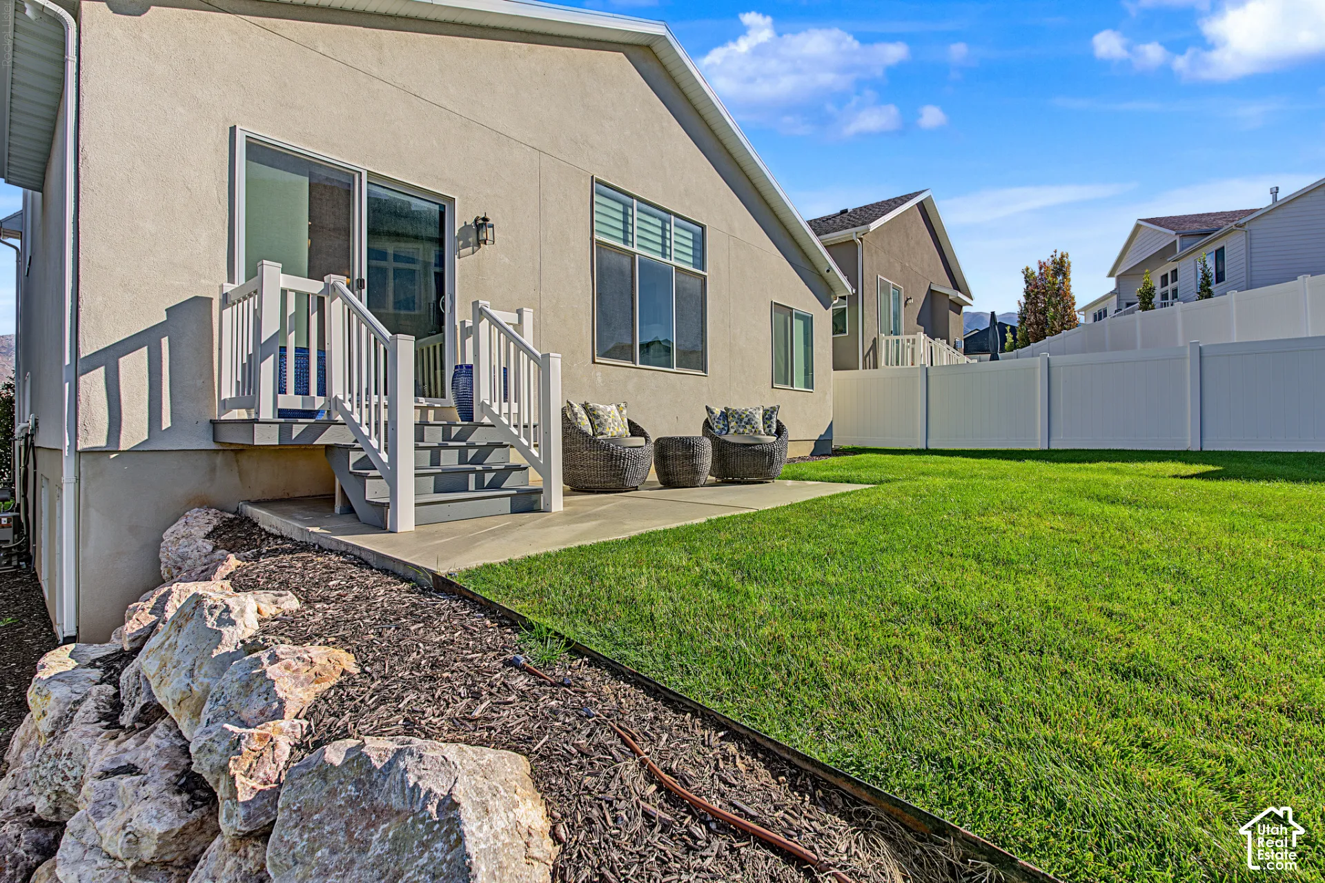 Rear exterior featuring a concrete patio, white vinyl privacy fencing, tiered landscaping with stone retaining wall, and sliding glass door with stairs