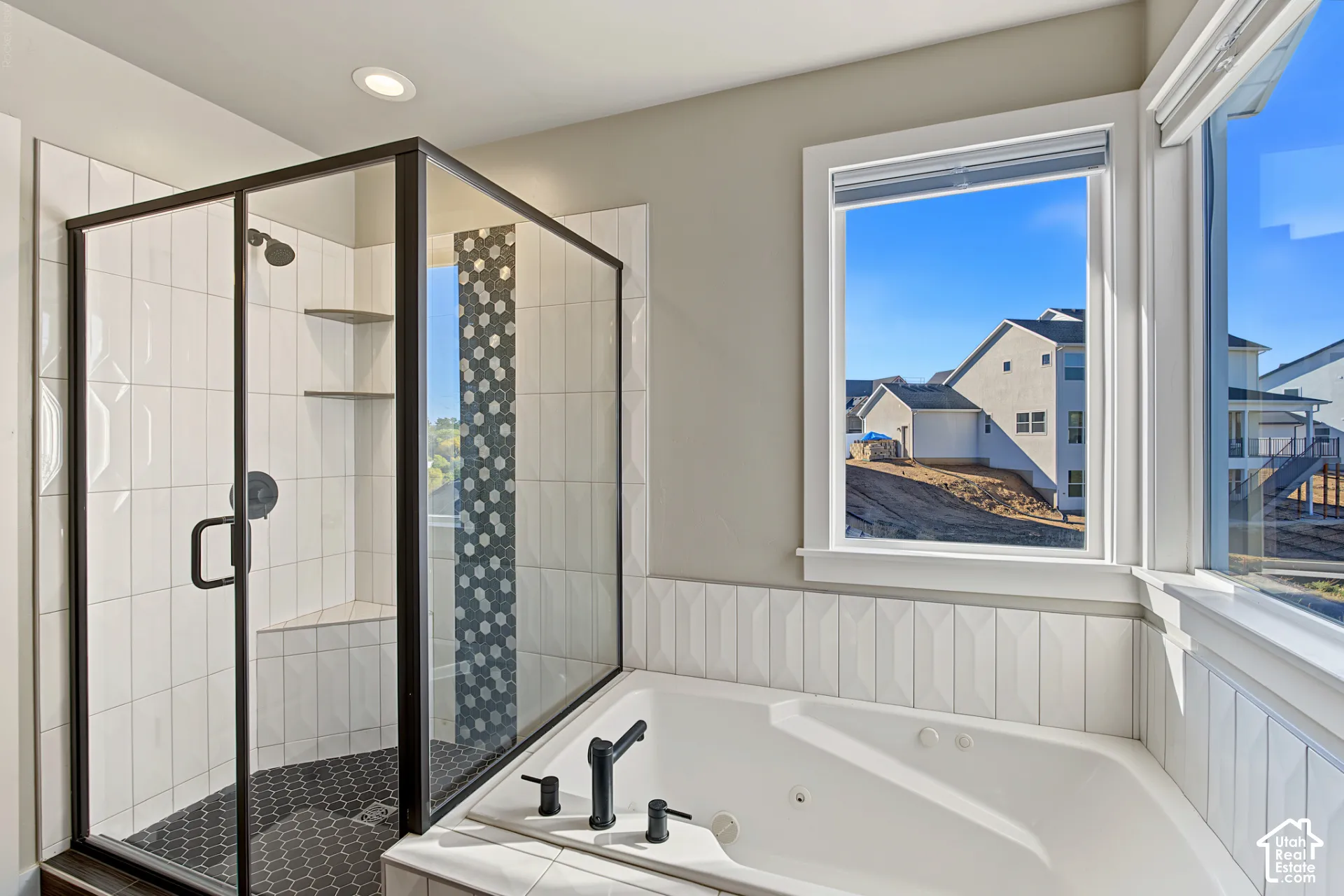 Walk-in shower with dark-framed glass enclosure, built-in shelving, and mosaic floor tile, accompanied by a jetted soaking tub and textured white wall tile