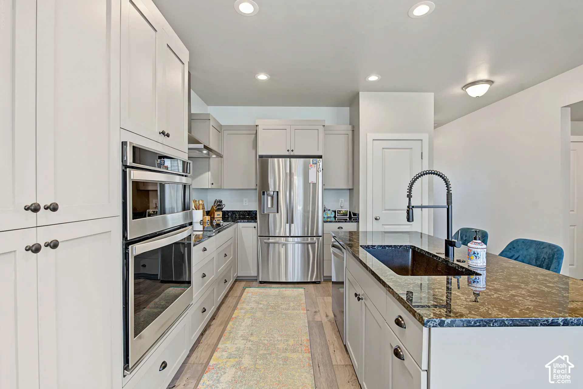 Kitchen featuring light-toned cabinetry, stainless steel double wall ovens and refrigerator, stone-surfaced island countertop, undermount sink with a matte black pull-down faucet, and wood-finish flooring