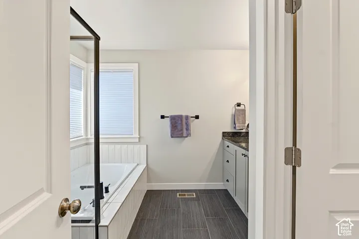 Built-in soaking tub with a white vertical panel surround, dark tile flooring, vanity cabinet with a dark stone-finish countertop, and matte black hardware fixtures