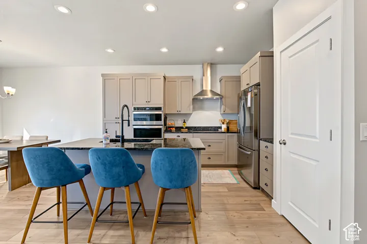 Kitchen featuring Shaker-style cabinetry, a central island with a stone countertop and dark finish faucet, stainless steel double wall ovens, a chimney-style range hood, and wood-finish flooring