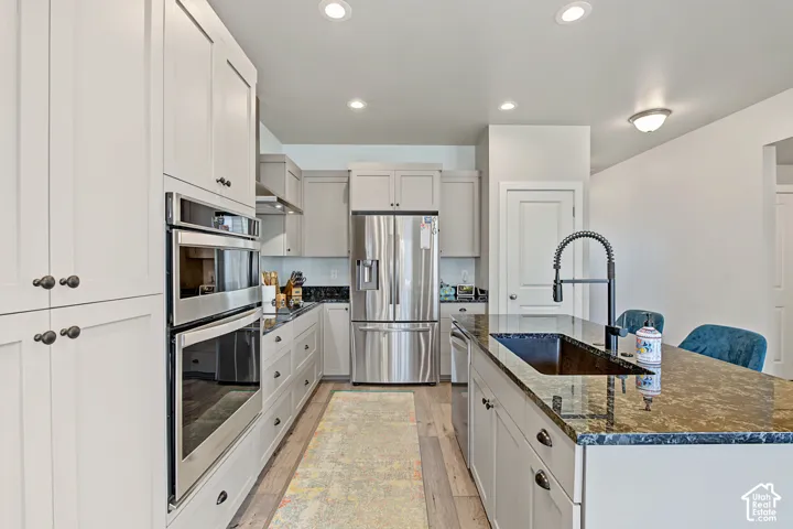 Kitchen featuring light-toned cabinetry, stainless steel double wall ovens and refrigerator, stone-surfaced island countertop, undermount sink with a matte black pull-down faucet, and wood-finish flooring