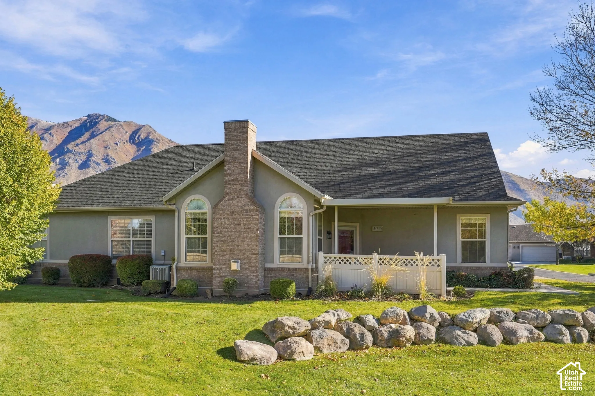Single-story residence featuring a partial stone facade, prominent chimney, covered front porch with lattice railing, and mountain backdrop