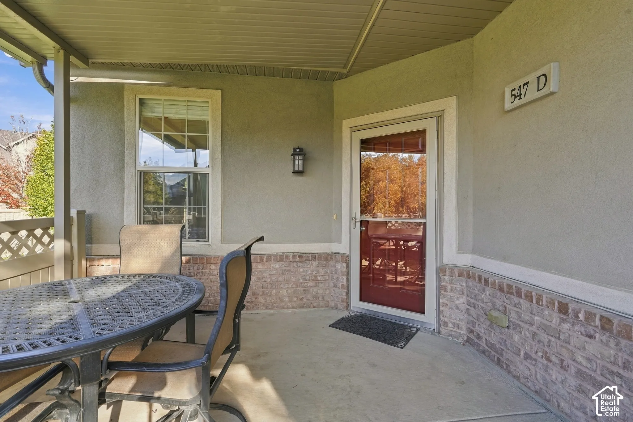 Covered patio featuring concrete flooring, a masonry brick base, stucco walls, a glass storm door, and an exterior wall-mounted light fixture