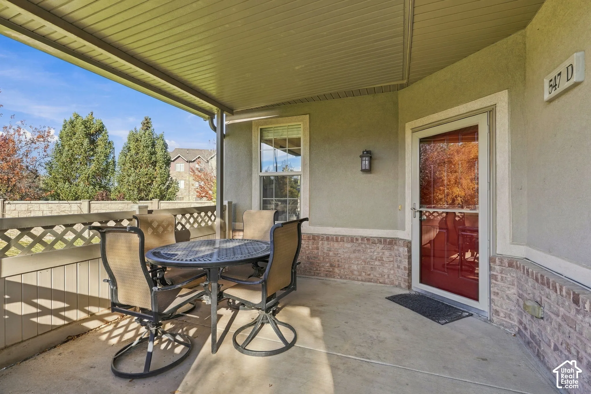 Covered patio featuring concrete flooring, partial brick exterior wall, lattice-style perimeter railing, and wall-mounted exterior lighting