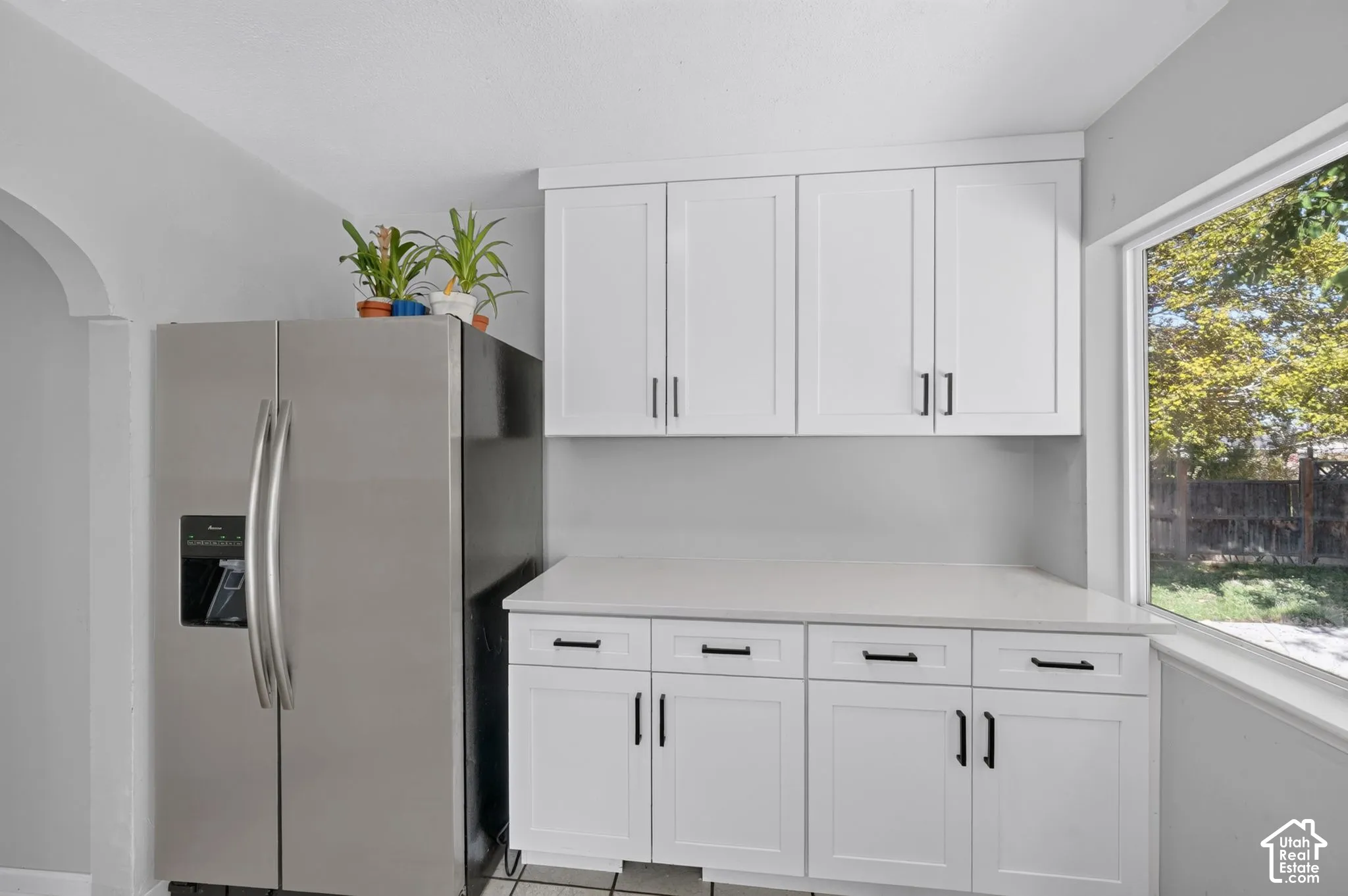 Shaker-style white cabinetry with matte black hardware, stone-composite countertop, stainless steel side-by-side refrigerator, and large window with natural lighting