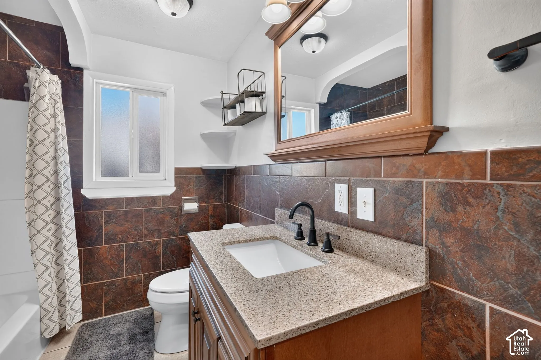 Bathroom vanity featuring a stone-composite countertop, undermount sink, and matte black widespread faucet, complemented by full-height tile wall surrounds and a built-in wood-frame mirror