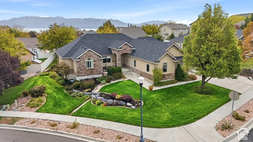 Single-story residence featuring stone and stucco exterior, multi-gabled shingle roof, concrete driveway, and professionally landscaped yard with tiered rock garden beds