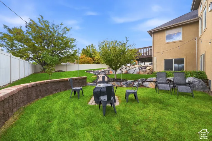 Backyard featuring a tiered stone retaining wall, paved fire pit area, elevated wood deck, and white vinyl perimeter fencing