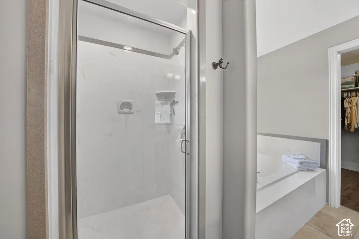 Walk-in shower featuring glass enclosure, wall-mounted metallic fixtures, and light-toned stone-finish surround, accompanied by a soaking tub with matching stone-finish deck