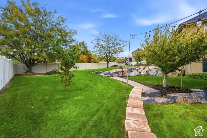 Backyard featuring a tiered stone retaining wall, paved stone pathway, mature trees, manicured lawn, and a vinyl privacy fence