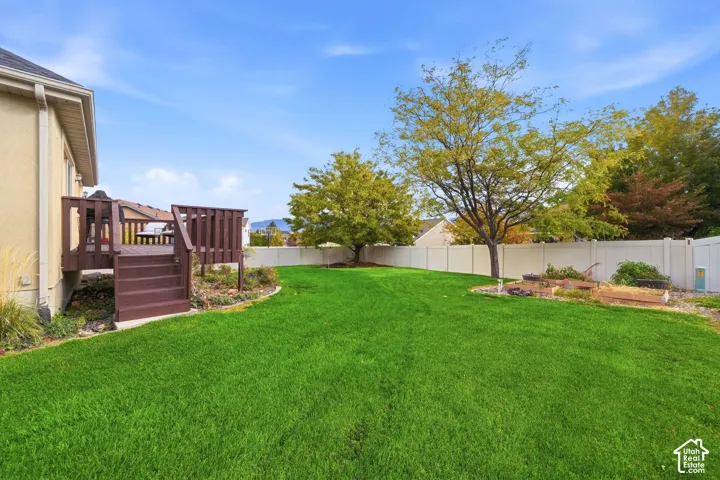 Backyard featuring a raised wood deck, expansive lawn, white vinyl fencing, mature trees, and garden beds with stone edging