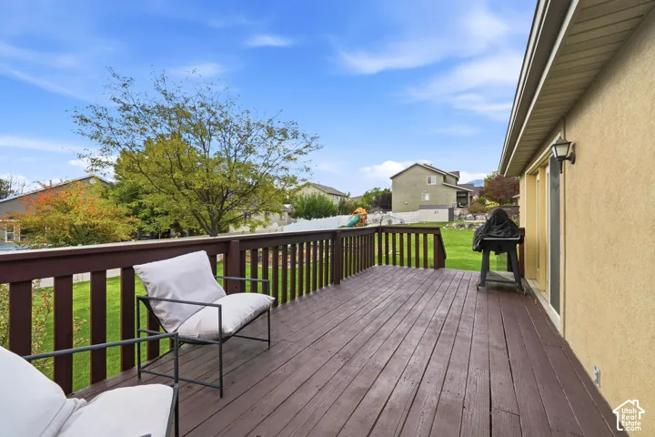 Elevated deck featuring wood-finish decking and a wooden railing, overlooking a lawn with mature trees and a vinyl perimeter fence