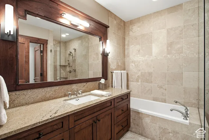 Bathroom featuring a custom wood vanity with a stone countertop, an integrated undermount sink, a full-width mirror with a decorative wood frame, wall-mounted light fixtures, and a tub-shower combination surrounded by natural stone tile