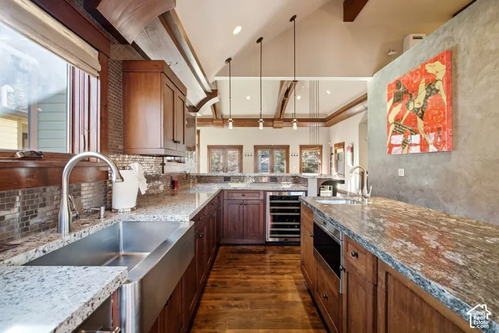 Kitchen featuring wood cabinetry, stone countertops, a stainless steel apron-front sink, integrated wine refrigeration, and a vaulted ceiling with exposed structural beams