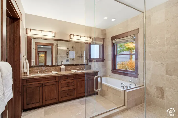 Dual-sink vanity with stained wood cabinetry, stone countertops, frameless glass-enclosed shower, tile-surround soaking tub, and recessed lighting