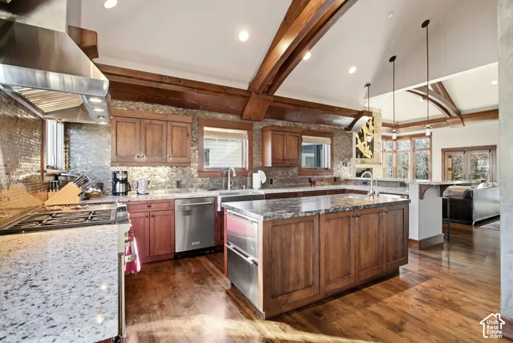 Kitchen featuring vaulted ceilings with exposed wood beams, wood-finish flooring, stone-tiled backsplash, expansive stone-topped center island, and stainless steel appliances