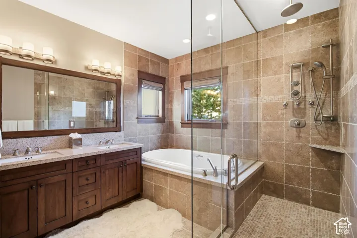 Bathroom featuring a double vanity with stone countertop, wood cabinetry, stone-tiled walls, glass-enclosed shower, and a built-in soaking tub