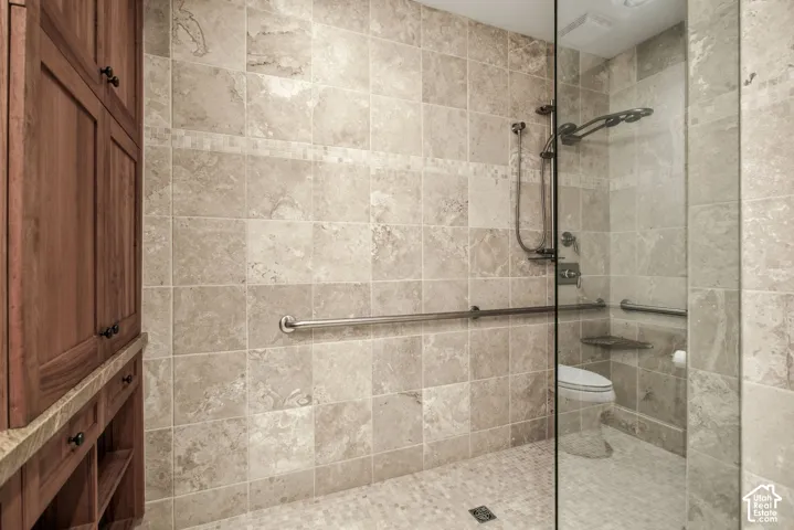 Walk-in shower featuring floor-to-ceiling stone tile, a glass partition, adjustable shower head, built-in corner shelving, and stainless steel grab bars, complemented by wooden built-in cabinetry with stone surfacing