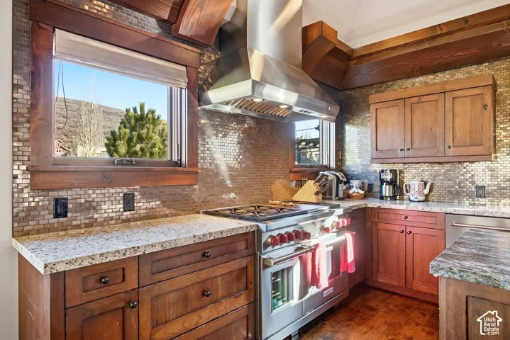 Kitchen featuring custom wood cabinetry, stone countertops, stainless steel professional-grade range and chimney hood, and metallic mosaic tile backsplash, framed by exposed wood ceiling beams