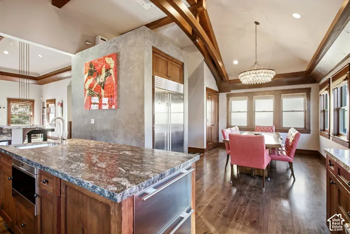Vaulted ceilings with exposed timber beams, natural wood-finish flooring, custom wood cabinetry, stone-surface kitchen island, and a decorative crystal chandelier