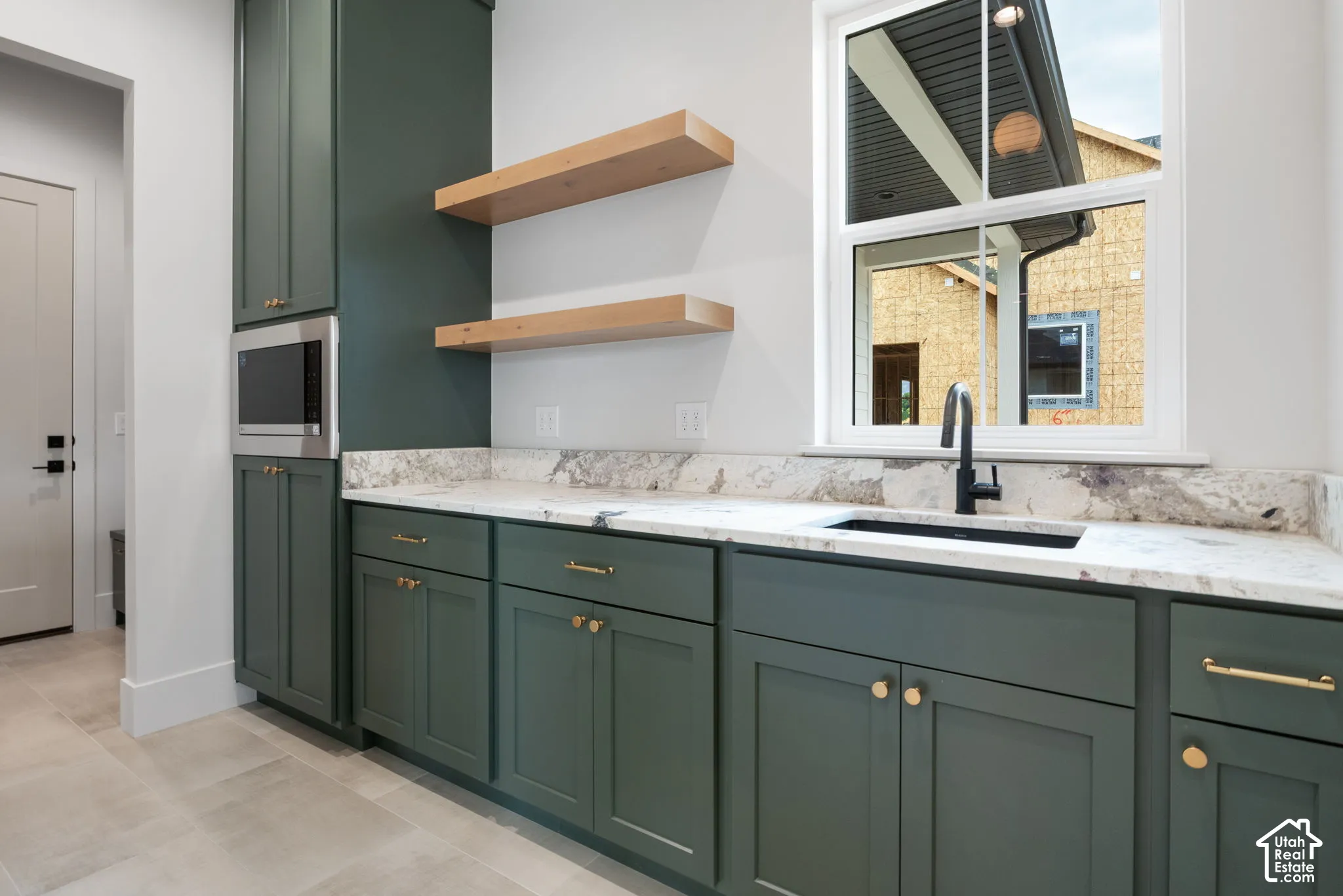 Deep green cabinetry featuring brass-tone hardware, stone countertops, integrated microwave, natural wood floating shelves, and a matte black faucet with an under-mount sink