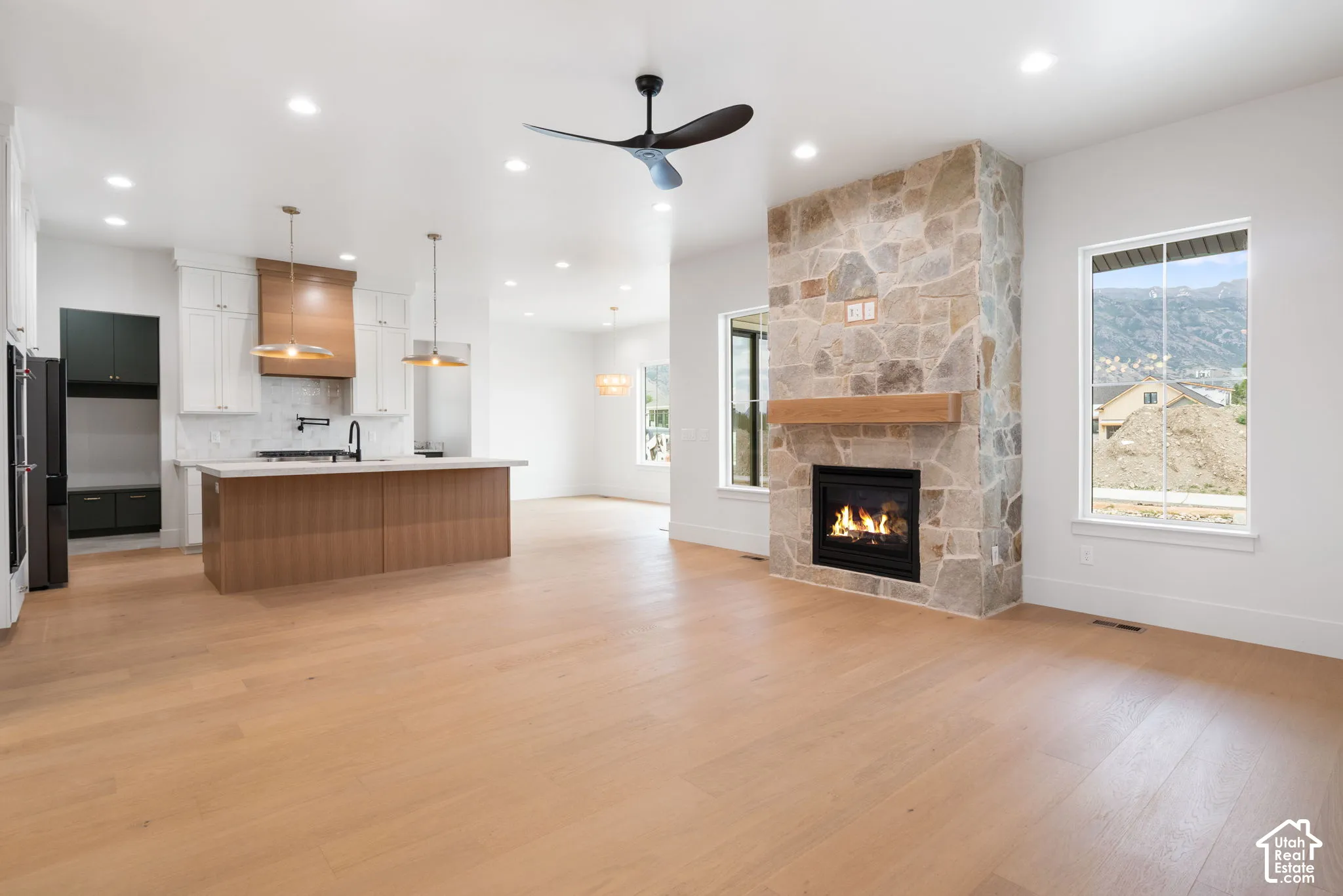 Open-concept interior featuring light wood-finish flooring, a full-height stone fireplace with a wood mantel, an island kitchen with white cabinetry, and recessed lighting