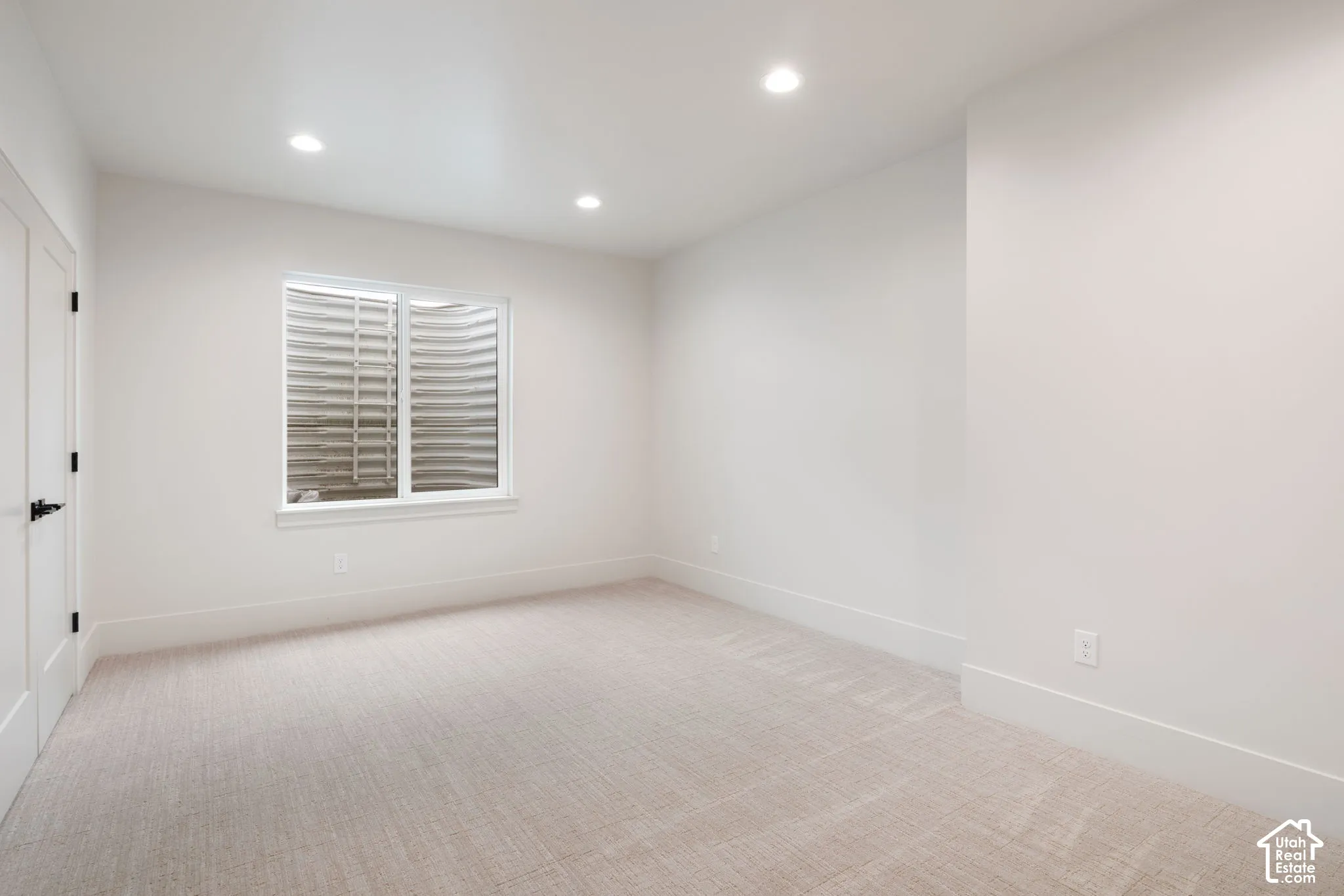 Neutral-toned interior space featuring wall-to-wall carpeting, recessed ceiling lighting, and an egress window with a corrugated metal window well