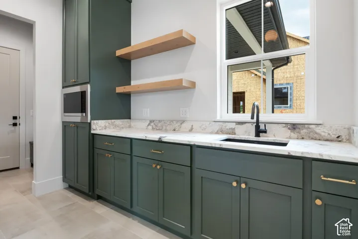 Deep green cabinetry featuring brass-tone hardware, stone countertops, integrated microwave, natural wood floating shelves, and a matte black faucet with an under-mount sink