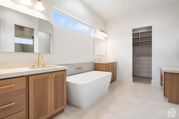 Freestanding soaking tub with a wall-mounted brass faucet, wood-finish cabinetry, neutral stone-finish countertops, arched mirrors, and a walk-in closet featuring built-in shelving