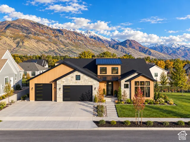 Contemporary residence featuring a multi-textured exterior of stone veneer and vertical wood-finish siding, black metal roofing, two attached garages with horizontal glass panels, and a backdrop of mountain scenery