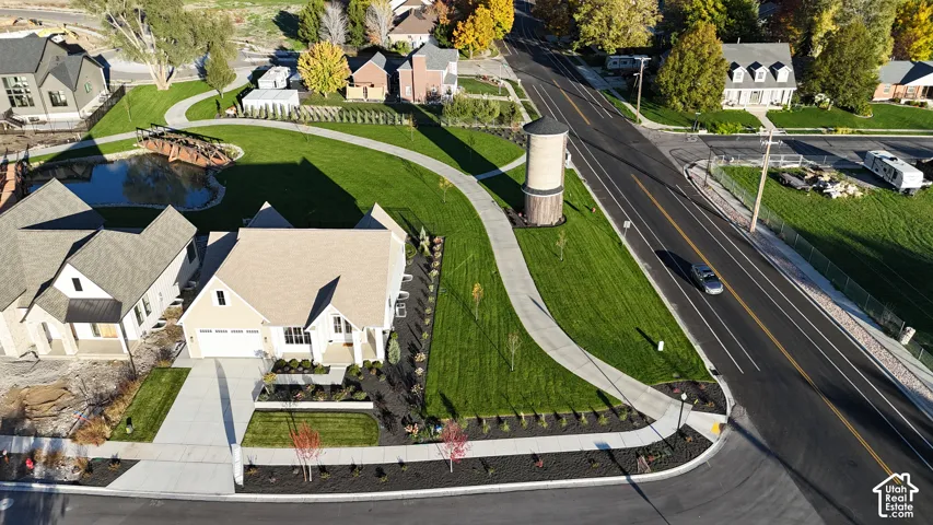 Aerial view of a corner lot property featuring an extensive manicured lawn, paved pedestrian walkway, water feature with a wooden footbridge, and a prominent water tower structure