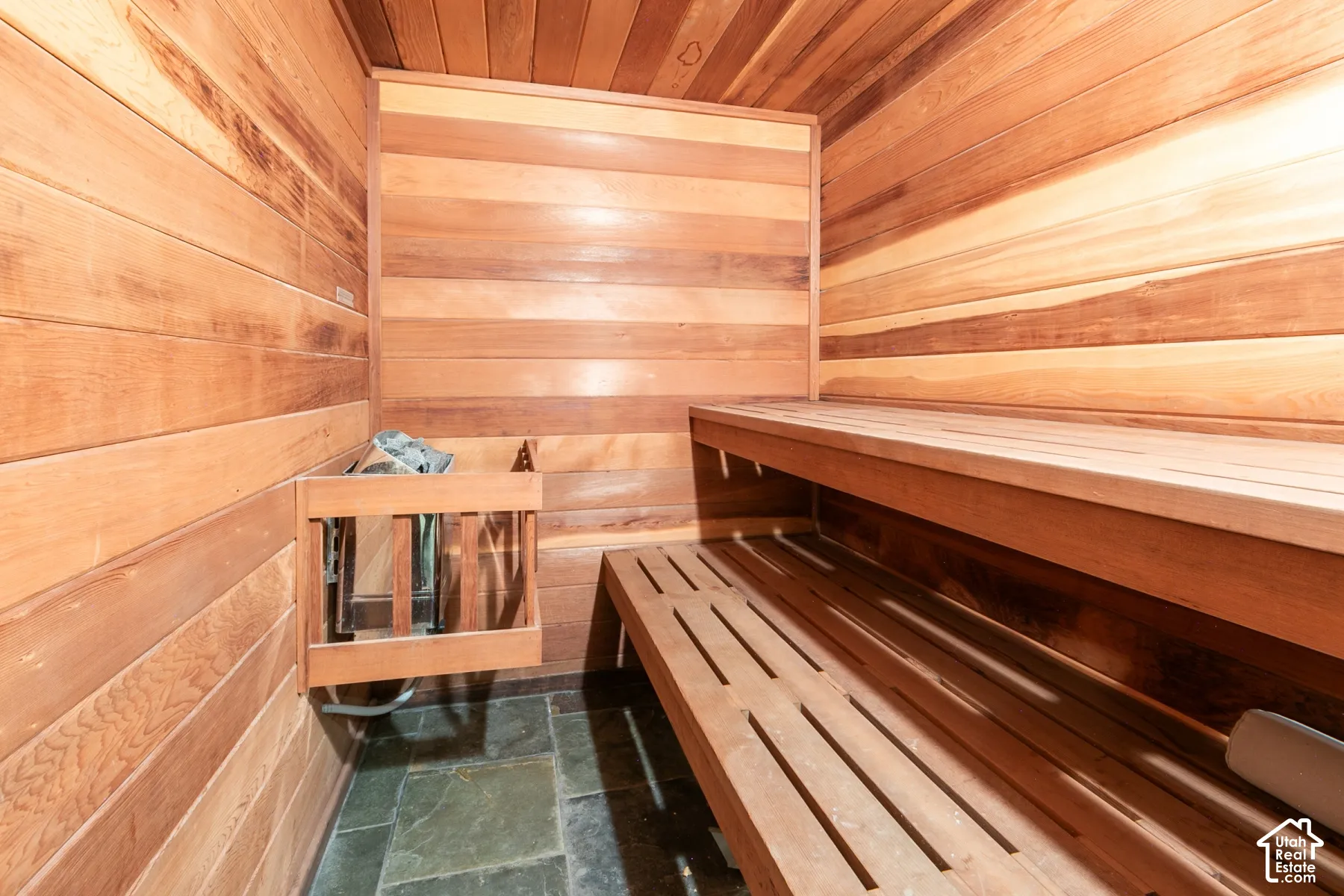 Sauna featuring horizontal wood-paneled walls and ceiling, multi-level slatted wood bench seating, and stone tile flooring