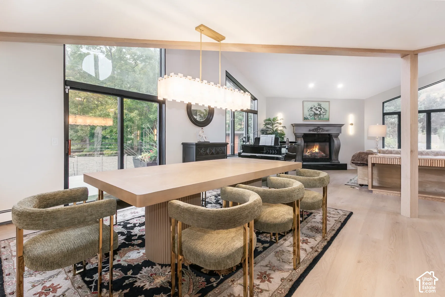 Open-concept living space featuring wood-finish flooring, floor-to-ceiling windows with black frames, a contemporary linear chandelier, and a masonry fireplace